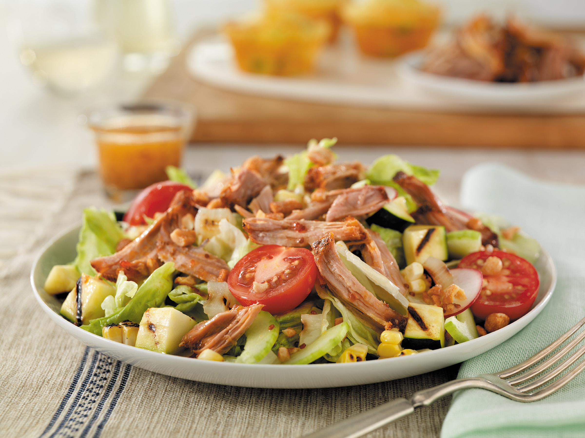 A colorful salad bowl filled with fresh greens, topped with tender pulled pork that has been cooked low and slow until it's melt-in-your-mouth juicy and flavorful. The salad is also garnished with sliced cherry tomatoes, fresh cucumbers, grilled zucchini and corn.